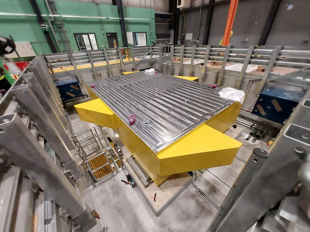 Wide angle view of the yellow painted shaking table with machined surface in position within its pit.
